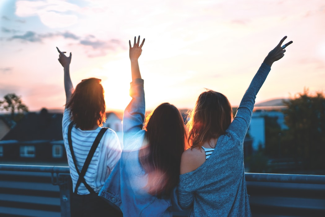trois jeunes femmes de dos levant les mains heureuses face à un levé de soleil