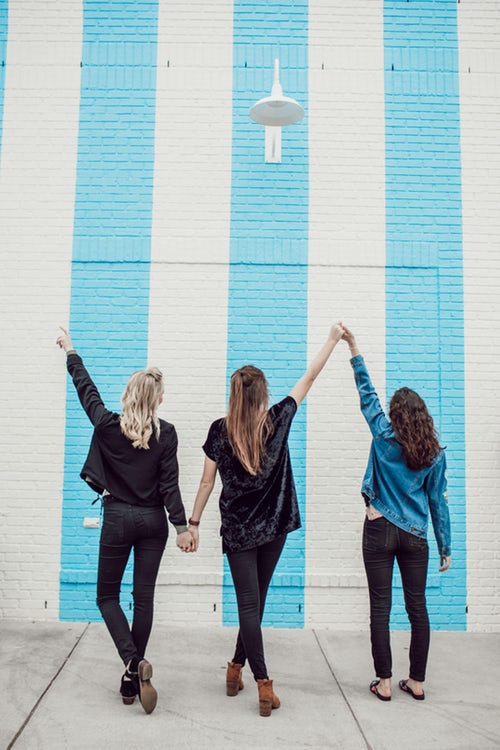 trois jeune femmes de dos se tenant la main devant un mur couleur blanc et bleu